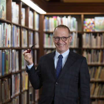A photograph of David Sedaris stood in a library, he is holding a pipe and wearing a blue blazer, shirt and tie, he is grinning in to the camera