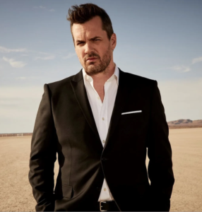 A photo of comedian Jim Jefferies wearing a black suit and white shirt stood in a desert