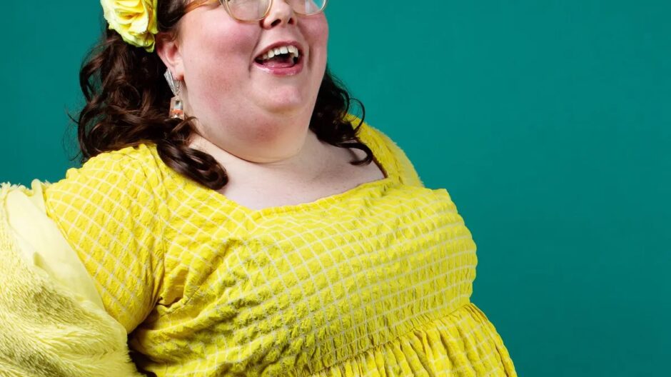 A photo of comedian Alison Spittle. She has curly shoulder length brown hair and is stood in front of a green background wearing a yellow dress and matching flower crown
