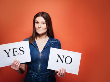 A photo of comedian and actor Fiona Allen, she is stood in front of an orange background and she has long dark hair. She is wearing a denim jumpsuit and is holding two pieces of paper one saying yes and the other saying no