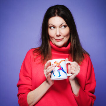Comedian and actor Fiona Allen stood in front of a purple background, wearing a red jumper and holding a mug