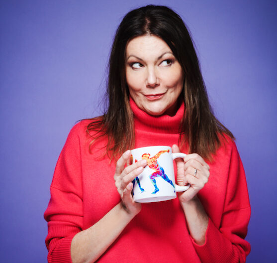 Comedian and actor Fiona Allen stood in front of a purple background, wearing a red jumper and holding a mug