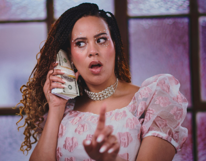 A photograph of comedian Mary O'Connell, she is wearing a white and pink dress and holding a wad of cash up to her ear like its a phone