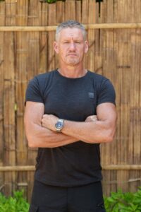 A photo of Billy Billingham, he has short grey hair and is wearing a black t shirt, he has his arms crossed and is looking directly in to the camera